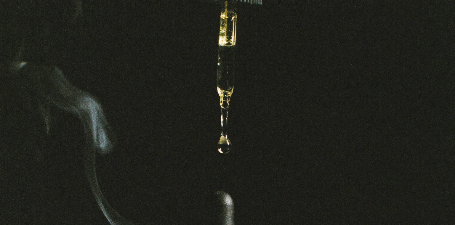 SÉVE Layering Oil — an amber dropper bottle on dark stone, a single drop falling.